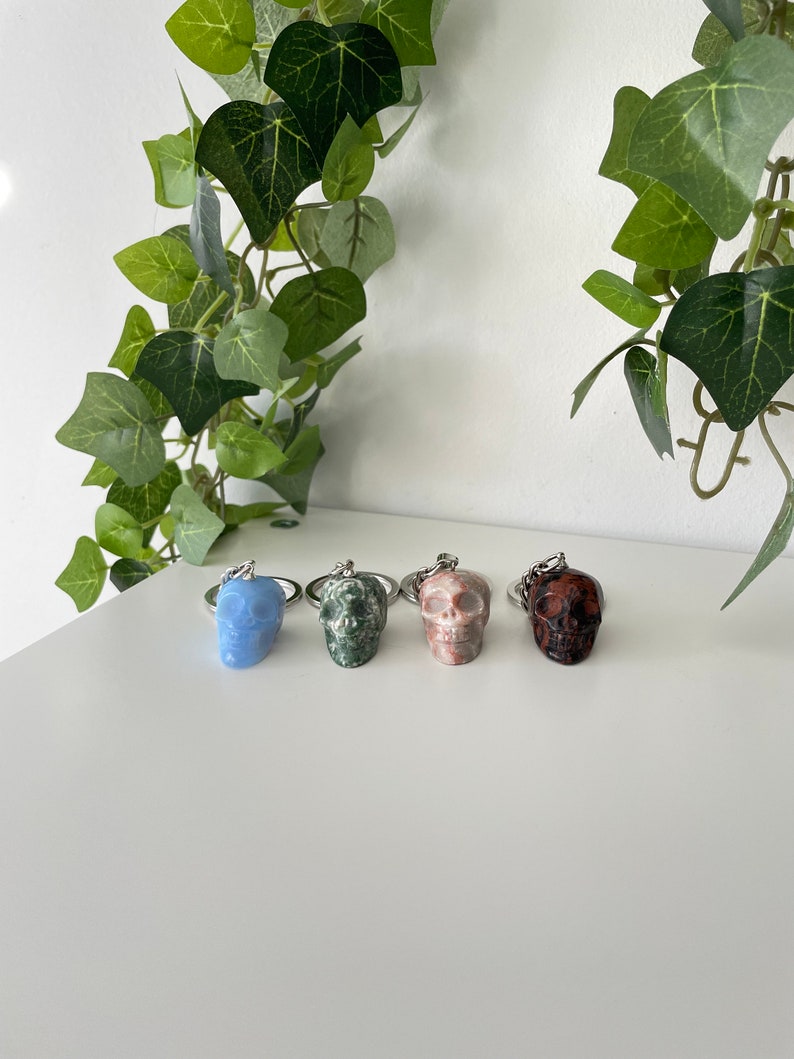 Puede incluir: Cuatro llaveros con forma de calavera hechos de diferentes tipos de piedra. Los llaveros est&aacute;n dispuestos en una fila de izquierda a derecha: azul claro, verde, rosa y rojo oscuro.