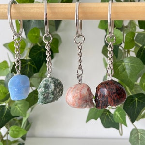 Puede incluir: Cuatro llaveros con forma de calavera hechos de diferentes tipos de piedra. Los llaveros est&aacute;n colgados de una varilla de madera. Las calaveras son azules, verdes, rosas y marrones.
