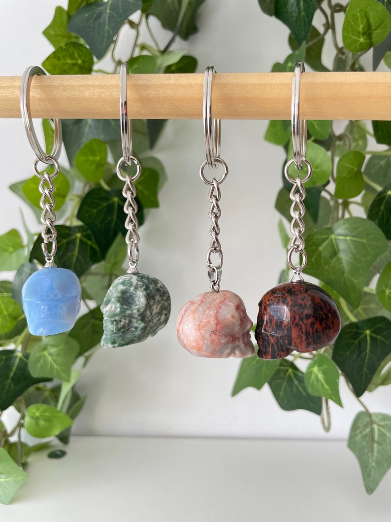 Puede incluir: Cuatro llaveros con forma de calavera hechos de diferentes tipos de piedra. Los llaveros est&aacute;n colgados de una varilla de madera. Las calaveras son azules, verdes, rosas y marrones.