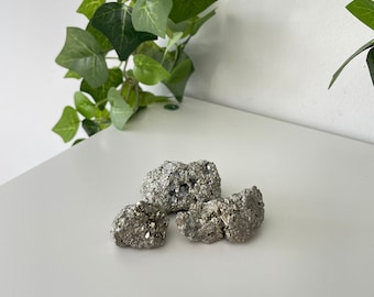 May include: Three pieces of raw pyrite crystals, also known as fool's gold, on a white surface. The crystals are a silvery-gold color and have a rough, textured surface.