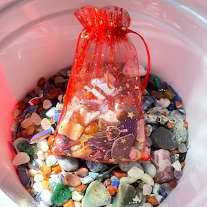 May include: A white bucket filled with a variety of colorful stones and crystals. A red mesh bag with gold stars is filled with more stones and crystals and sits on top of the stones in the bucket.