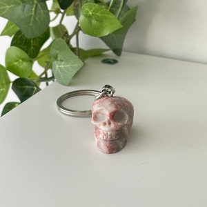 Puede incluir: Un peque&ntilde;o llavero de calavera tallado en m&aacute;rmol rosa y blanco. La calavera tiene una cara detallada con cuencas de los ojos, una nariz y una boca.