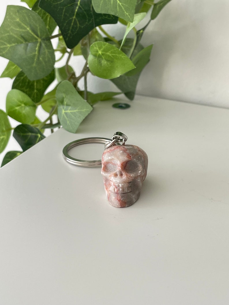 Puede incluir: Un peque&ntilde;o llavero de calavera tallado en m&aacute;rmol rosa y blanco. La calavera tiene una cara detallada con cuencas de los ojos, una nariz y una boca.