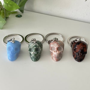 Puede incluir: Cuatro llaveros con forma de calavera hechos de diferentes tipos de piedra. Las calaveras son azules, verdes, rosas y marrones. Cada llavero tiene un anillo y una cadena plateados.