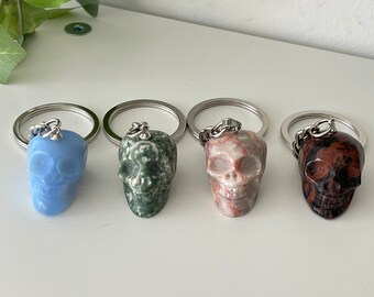 May include: Four skull-shaped keychains made of different types of stone. The skulls are blue, green, pink, and brown. Each keychain has a silver ring and chain.