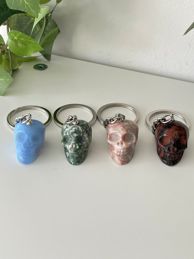 Puede incluir: Cuatro llaveros con forma de calavera hechos de diferentes tipos de piedra. Las calaveras son azules, verdes, rosas y marrones. Cada llavero tiene un anillo y una cadena plateados.