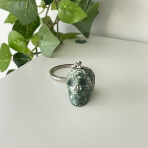 Puede incluir: Llavero de calavera de piedra verde y blanca. La calavera est&aacute; tallada en una piedra verde y blanca con un anillo plateado.