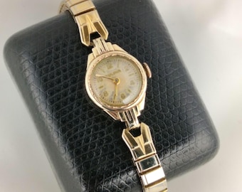 May include: A vintage Bulova wristwatch with a gold-tone case and band. The watch face is cream-colored with gold hands and numerals. The watch is displayed on a black, textured surface.