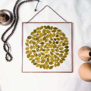May include: Square copper-framed artwork featuring a circular arrangement of green leaves. The leaves are arranged on a white background. A wooden beaded necklace and two wooden decorative objects are also present.