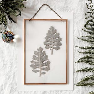 May include: A framed botanical art piece featuring two gray, textured leaves. The leaves are displayed on a white background within a copper-colored metal frame, suspended by a chain. Ferns and a decorative glass ball surround the frame.