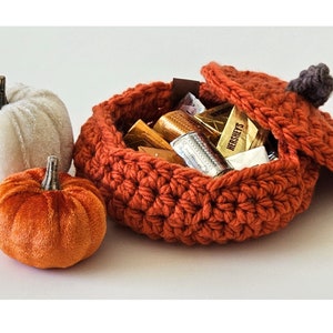 Puede incluir: Una cesta de calabaza de ganchillo con tapa, llena de caramelos de chocolate surtidos. La cesta es naranja y la tapa tiene un asa de tela marrón. Hay dos pequeñas calabazas, una blanca y otra naranja, en el fondo.