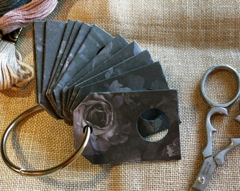 May include: A set of black and white fabric swatches with a floral design, attached to a silver ring. The swatches are arranged in a fan shape. A pair of silver embroidery scissors are also in the image.