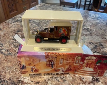 May include: A brown 1927 Talbot South Pacific delivery truck model with a white roof and a red and white South Pacific Lager logo on the side. The model is in a clear display case on a brown and white marble surface. The box for the model is visible in the background and features a Matchbox logo and the text "Models of Yesteryear".