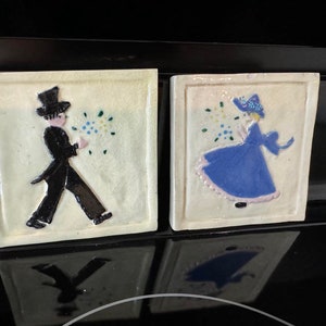May include: Two ceramic tiles with painted images of a man and a woman. The man is wearing a black suit and a top hat. The woman is wearing a blue dress and a white hat. Both figures are holding a bouquet of flowers.