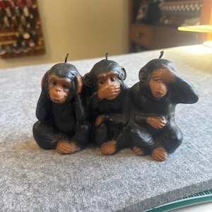 May include: A dark brown wax candle shaped as three chimpanzees sitting side by side. The first monkey covers its ears, the second covers its mouth, and the third covers its eyes. Each monkey has a small black wick protruding from the top of its head.