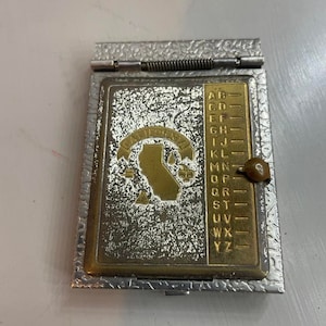 May include: A silver and gold metal compact with a California state outline and the word "California" in gold lettering. The compact has a built-in alphabet index on the right side.