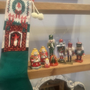 May include: A green and red knitted Christmas stocking with a white fireplace design hangs on a wooden shelf.  The shelf also displays a collection of small wooden nutcracker ornaments and ceramic angel figurines.
