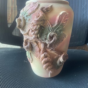 May include: A ceramic vase with a textured, floral design. The vase is decorated with raised, sculpted flowers and leaves in shades of green, brown, and pink.