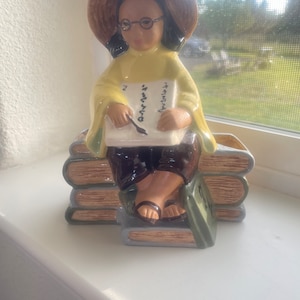 May include: A ceramic planter shaped like a girl wearing a yellow dress, a brown hat, and glasses, sitting on a stack of books. The girl is holding a book and a pen.