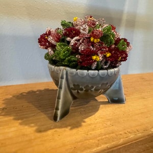 May include: A small ceramic planter with three legs, decorated with a floral pattern, holding a bouquet of red and pink beaded flowers.