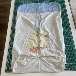 May include: White fabric baby diaper stacker with a light blue top and a duck applique. The stacker has two pockets and is secured with buttons. The duck applique features yellow bodies, pink feet, and black stitching.