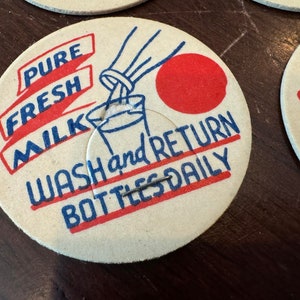 May include: A vintage milk bottle cap with a red and blue design. The cap says "Pure Fresh Milk" and "Wash and Return Bottles Daily".