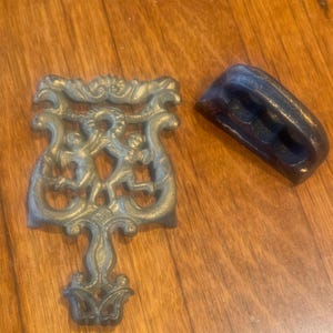 May include: Two antique cast iron trivets. One is a decorative trivet with a floral design and two figures. The other is a simple, rectangular trivet with three holes.