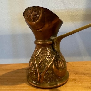 May include: A vintage copper and brass Turkish coffee pot with an ornate floral design. The pot has a long handle and a spout for pouring.