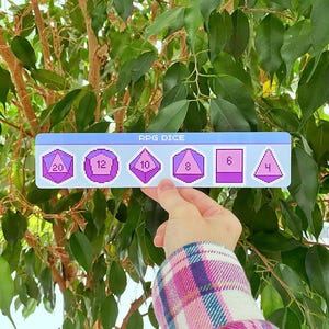 Könnte beinhalten: Ein rechteckiges Schild mit dem Text "RPG DICE" zeigt verschiedene Würfelformen, jeweils mit einer Zahl. Die Würfel sind in Lila- und Rosatönen gehalten und vor einem hellblauen Hintergrund platziert. Das Schild wird vor einer grünen Pflanze gehalten.