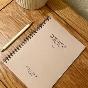 Può includere: Un quaderno beige a spirale con il testo "THINGS PEOPLE THINK I'VE GOT TIME FOR" e un piccolo simbolo di cuore. Il quaderno ha anche il testo "DAILY TO DO LIST" in basso.