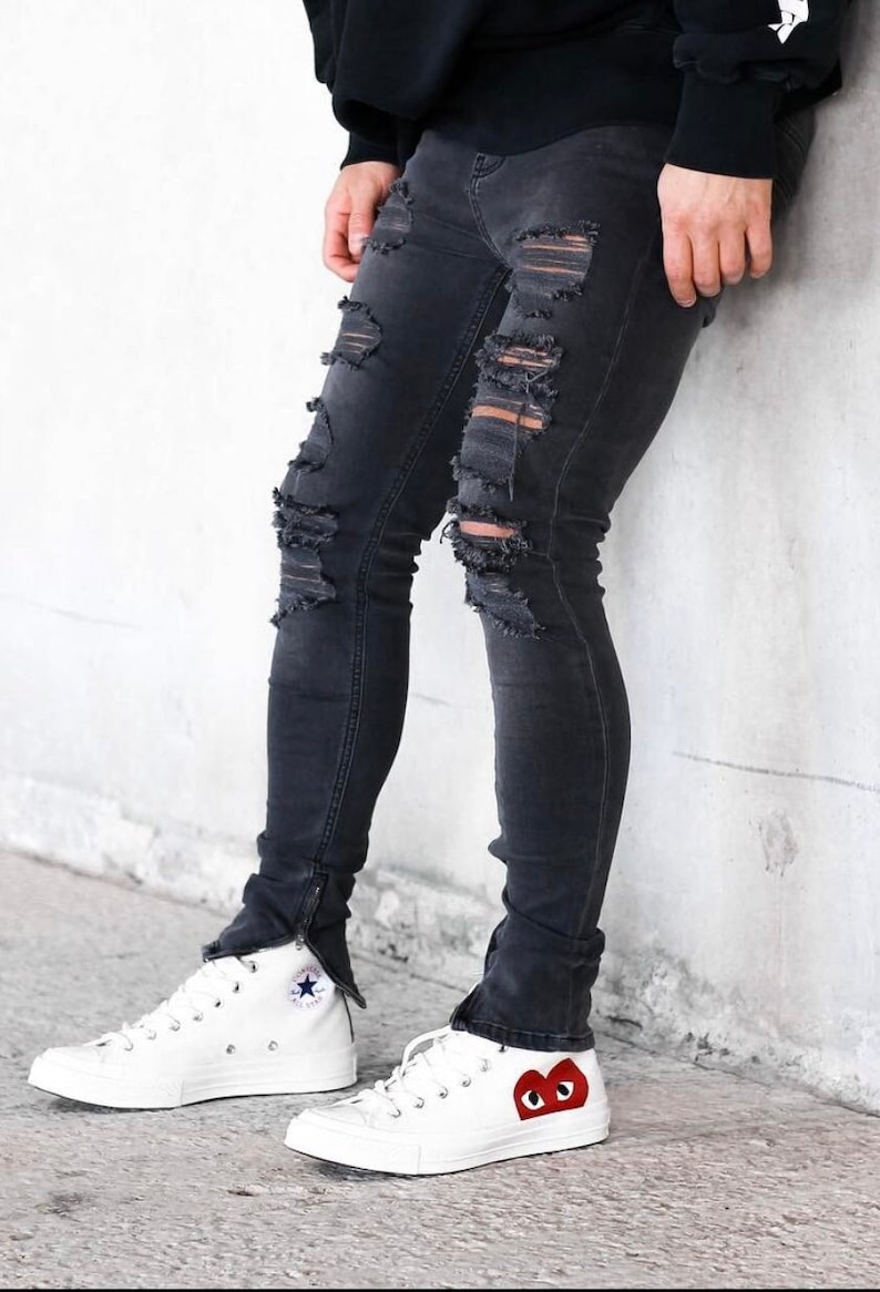 May include: A pair of white high-top sneakers with black trim and a red heart with eyes on one shoe. The sneakers are worn with black ripped jeans.