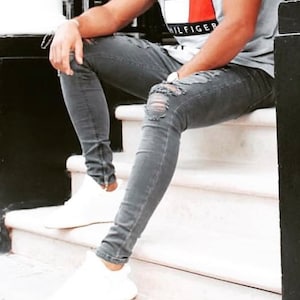 May include: A person wearing grey ripped jeans and white sneakers, sitting on a white step. The person is wearing a grey t-shirt with the Tommy Hilfiger logo in red, white, and blue.