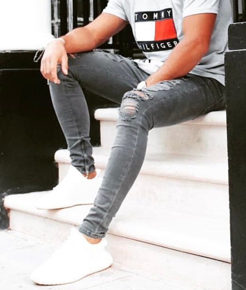 May include: A person wearing grey ripped jeans and white sneakers, sitting on a white step. The person is wearing a grey t-shirt with the Tommy Hilfiger logo in red, white, and blue.