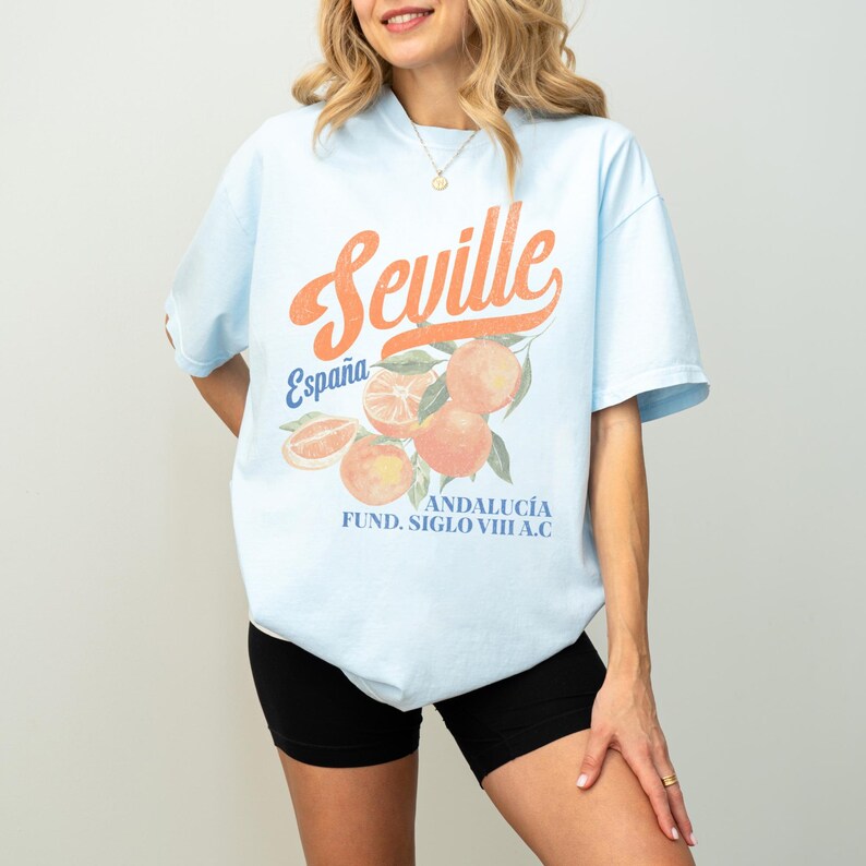 Pu&ograve; includere: Maglietta blu chiaro con una stampa grafica di arance e il testo "Sevilla Espa&ntilde;a Andalucia Fund. Siglo VIII A.C."