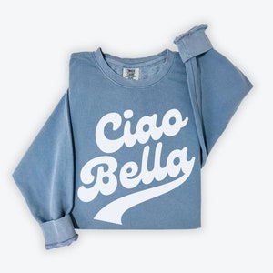 Puede incluir: Una sudadera azul con el texto "Ciao Bella" en blanco, con letras de estilo retro.