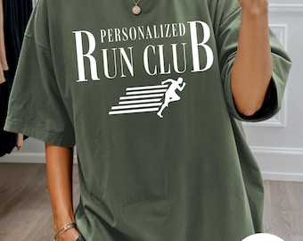 May include: An olive green crewneck t-shirt featuring the white text "PERSONALIZED RUN CLUB" above a graphic of a runner in motion with horizontal speed lines. The design is centred on the chest of the garment, which has short sleeves.