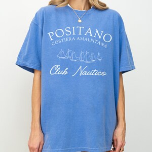 May include: A blue short-sleeve t-shirt featuring white screen-printed text that reads POSITANO COSTIERA AMALFITANA and Club Nautico, along with a graphic illustration of several sailboats positioned between the two lines of text.