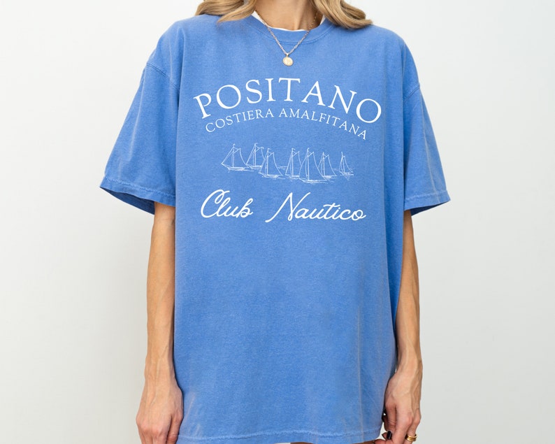 May include: A blue short-sleeve t-shirt featuring white screen-printed text that reads POSITANO COSTIERA AMALFITANA and Club Nautico, along with a graphic illustration of several sailboats positioned between the two lines of text.