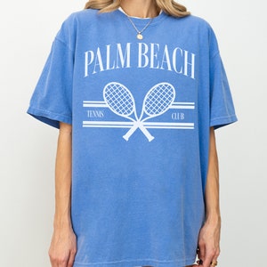 May include: An oversized blue t-shirt with a white graphic of two crossed tennis rackets and the text "PALM BEACH TENNIS CLUB".