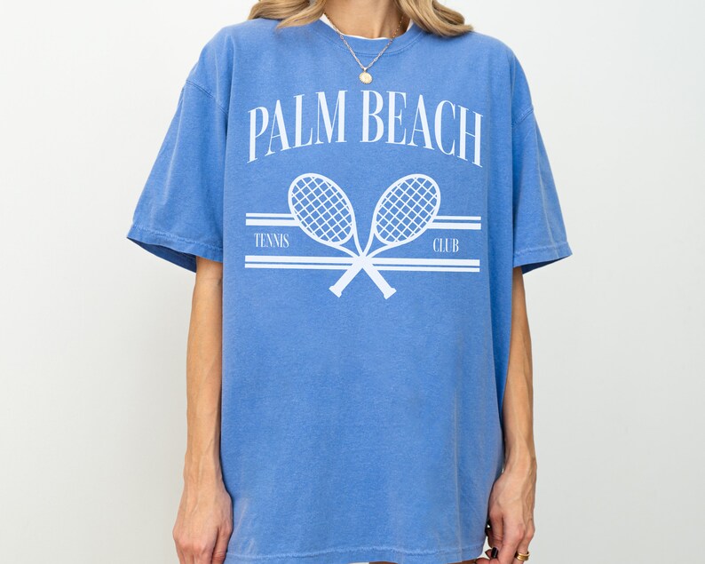 May include: An oversized blue t-shirt with a white graphic of two crossed tennis rackets and the text "PALM BEACH TENNIS CLUB".