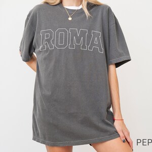 May include: A charcoal gray oversized t-shirt with a white outline of the word "ROMA" printed on the front.