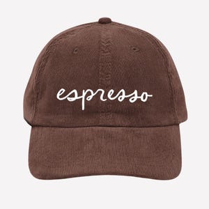 Peut inclure: Casquette de baseball en velours côtelé marron avec le mot "espresso" en écriture blanche sur le devant. La casquette a une visière incurvée et une couronne arrondie. Le chapeau est de couleur marron uni.