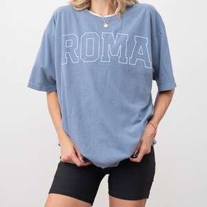 May include: A light blue oversized t-shirt with the word "ROMA" printed in white on the front. The shirt is made of a soft, comfortable fabric and has a relaxed fit.