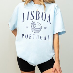 Puede incluir: Camiseta azul claro con un estampado gr&aacute;fico de una torre y el texto "LISBOA EST 1147 PORTUGAL" en azul marino.