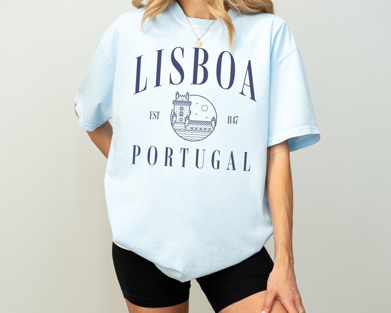 Puede incluir: Camiseta azul claro con un estampado gr&aacute;fico de una torre y el texto "LISBOA EST 1147 PORTUGAL" en azul marino.
