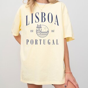 Puede incluir: Una camiseta amarilla con un dise&ntilde;o gr&aacute;fico azul que dice "LISBOA EST 1147 PORTUGAL" y presenta un dibujo de una torre y una luna.