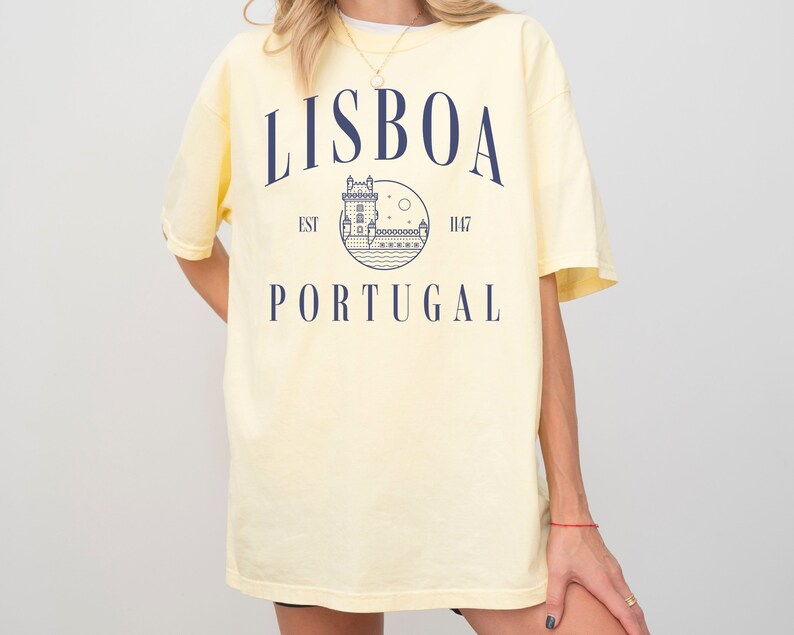 Puede incluir: Una camiseta amarilla con un dise&ntilde;o gr&aacute;fico azul que dice "LISBOA EST 1147 PORTUGAL" y presenta un dibujo de una torre y una luna.