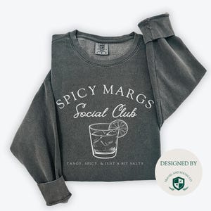 May include: A dark gray sweatshirt with the text "Spicy Margs Social Club" and a graphic of a margarita glass with a lime wedge. The text below the graphic reads "Tangy, Spicy, & Just a Bit Salty". The bottom right corner of the image shows a white circle with the text "Designed By Travel and Social Co."