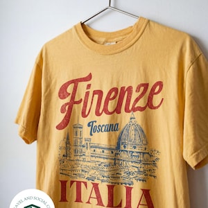 May include: Mustard yellow t-shirt with the word "Firenze" in red script above a blue and red illustration of a building. Below the illustration, the word "ITALIA" is printed in red. The shirt is hanging on a silver hanger.