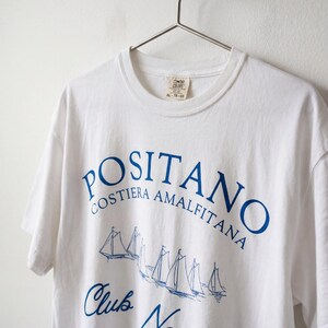 May include: A white short-sleeve t-shirt featuring blue screen-printed text that reads POSITANO COSTIERA AMALFITANA and Club Nautico, accompanied by a graphic illustration of several sailboats. The shirt has a crew neckline and a brand label.